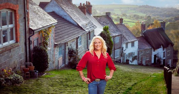 TV personality Sarah Beeney wearing a red shirt standing on a cobbled street with countryside in the background